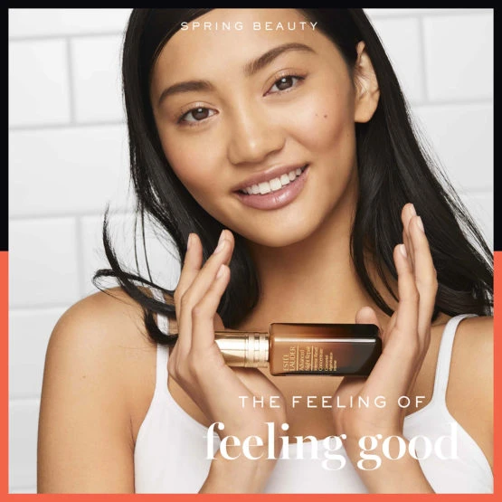 Smiling woman with tan skin and dark hair holds bottle of Estee Lauder Advanced Night Repair face serum in indoor beauty ad.