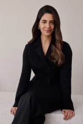 Portrait of a woman wearing a black double breasted blazer and trousers seated against a neutral wall, photographed by Amelia Dowd.