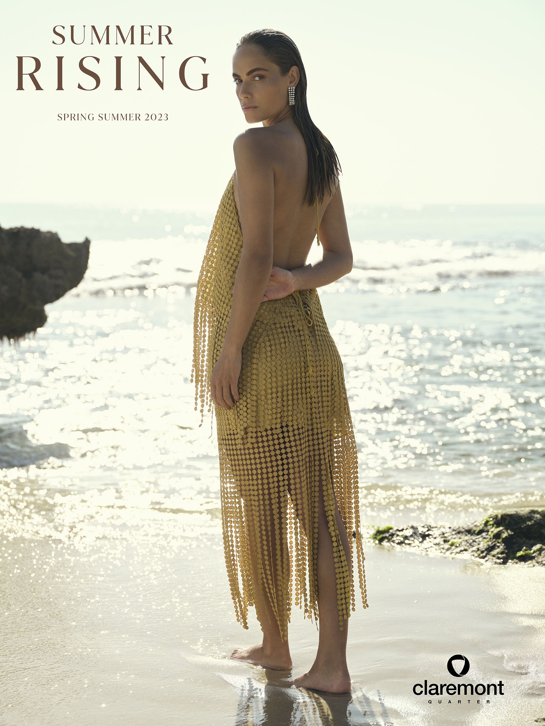 Fashion shot of a woman on a beach, wearing a gold, fringe dress, looking over her shoulder at the camera, soft, diffused lighting.