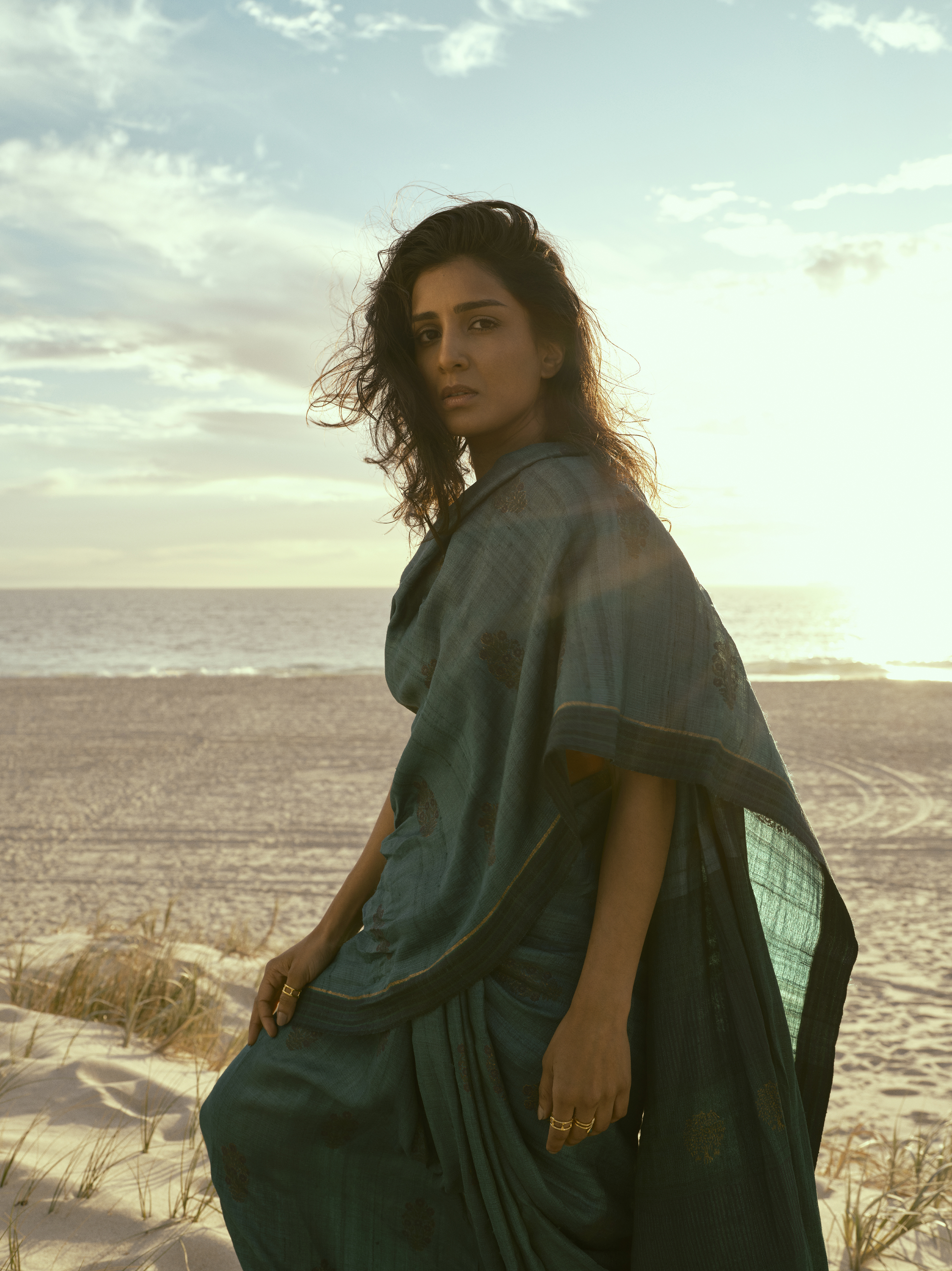Adult female in a teal sari poses on a sandy beach at sunset with the ocean and sky in the background, creating a serene fashion image.