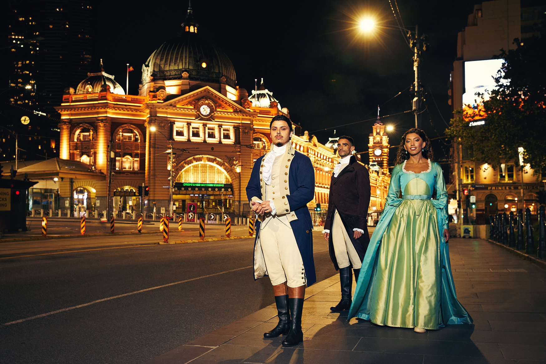 Three people in 18th-century costumes pose in front of Flinders Street Station in Melbourne at night, with bright city lights.