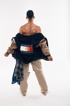 Back view of a muscular man with a jacket draped on his shoulders, wearing tan pants, captured against a bright white background.