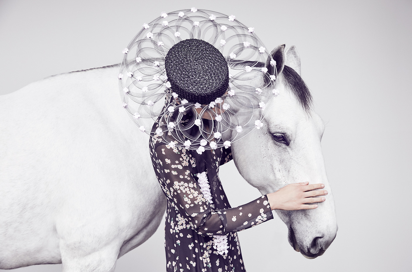 Fashion editorial featuring a woman in a floral dress and elaborate hat embracing a white horse against a light grey backdrop.