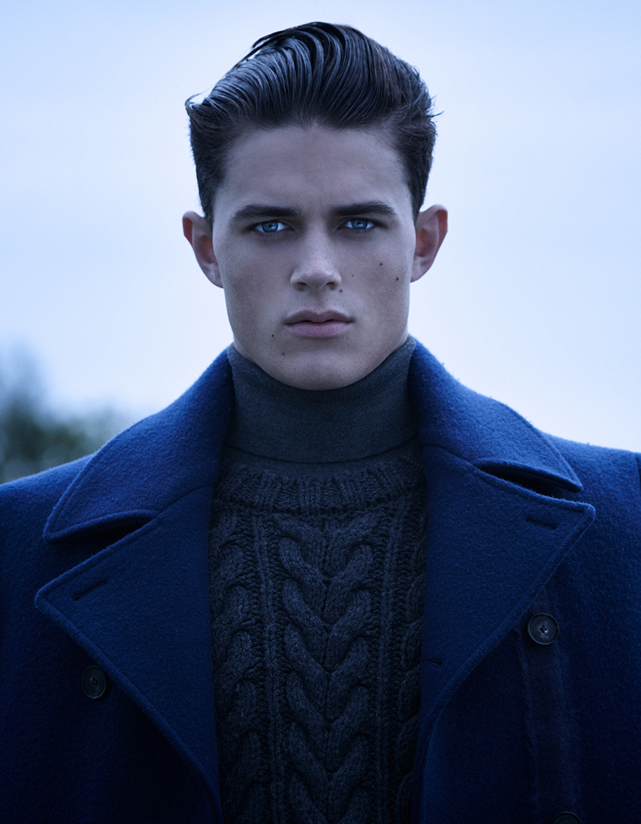 Portrait of a young adult male with slicked-back dark hair wearing a navy blue coat and gray sweater against a muted light-blue background.