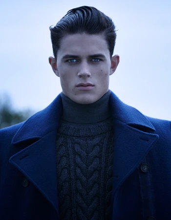 Portrait of a man with styled dark hair and blue eyes wearing a navy blue coat and a grey turtleneck sweater