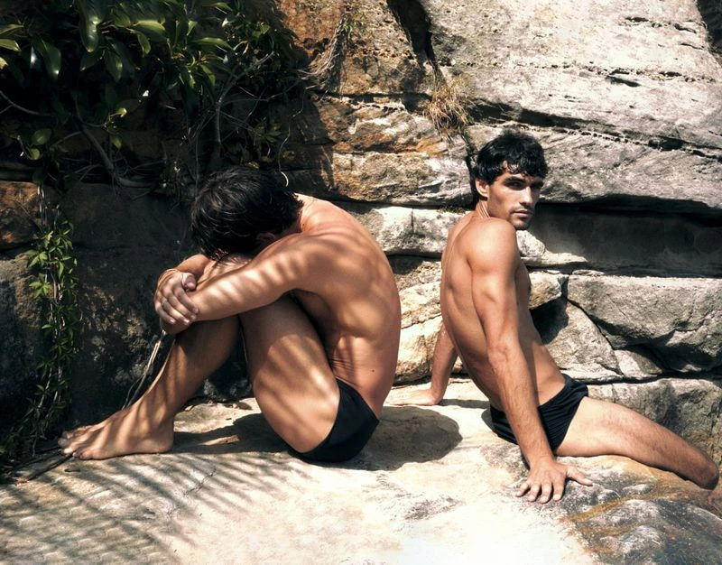 Two men in swimwear sit on rocks near green plants in sunlight. One man looks at the camera while the other rests his head on his knees.