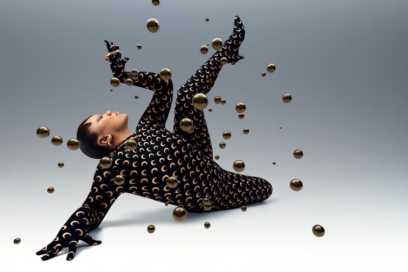Fashionable woman in crescent moon bodysuit, lying down with gold spheres surrounding her against a gradient gray background in a studio.