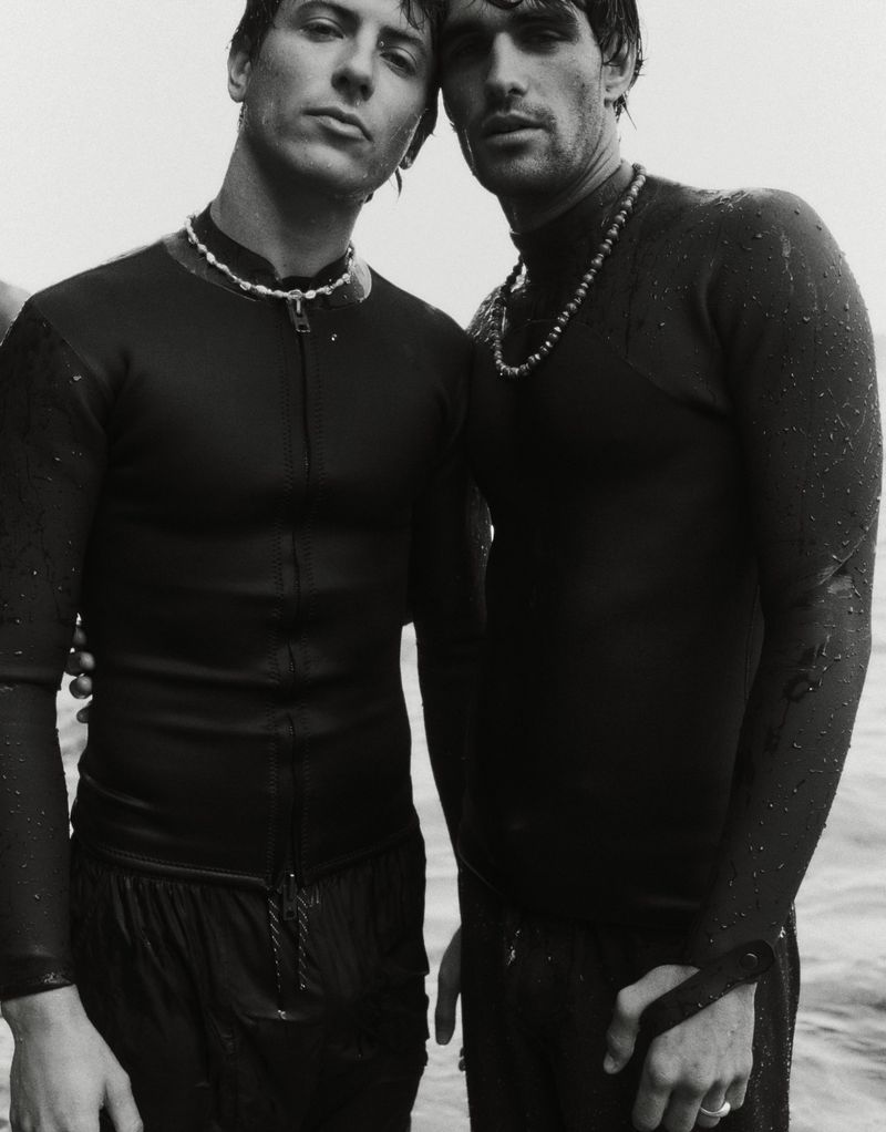 Monochrome portrait of two athletic adult men standing close together, wearing dark wetsuits and beaded necklaces near the ocean.