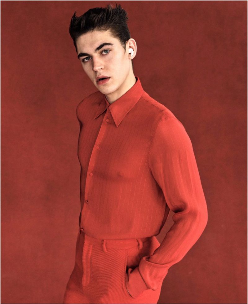 Portrait of a young adult male with short dark hair, wearing wireless earbuds and a red button-down shirt and pants against a matching red background.