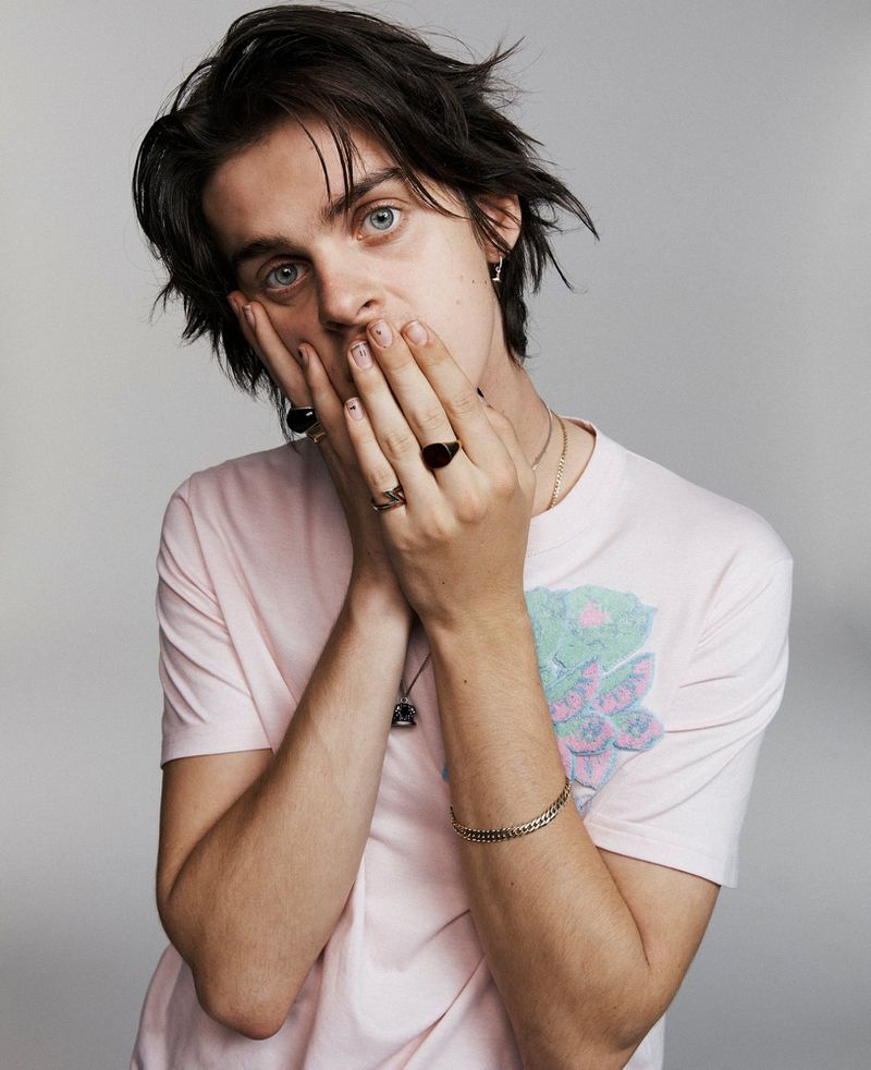 Portrait of a young adult with dark hair and blue eyes, wearing a pink t-shirt, with hands near the face, set against a grey background.