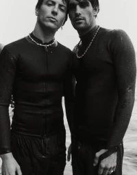 Two men wearing black wetsuits and beaded necklaces standing side by side in the water outdoors, styled by Mark Vassallo in Sydney.