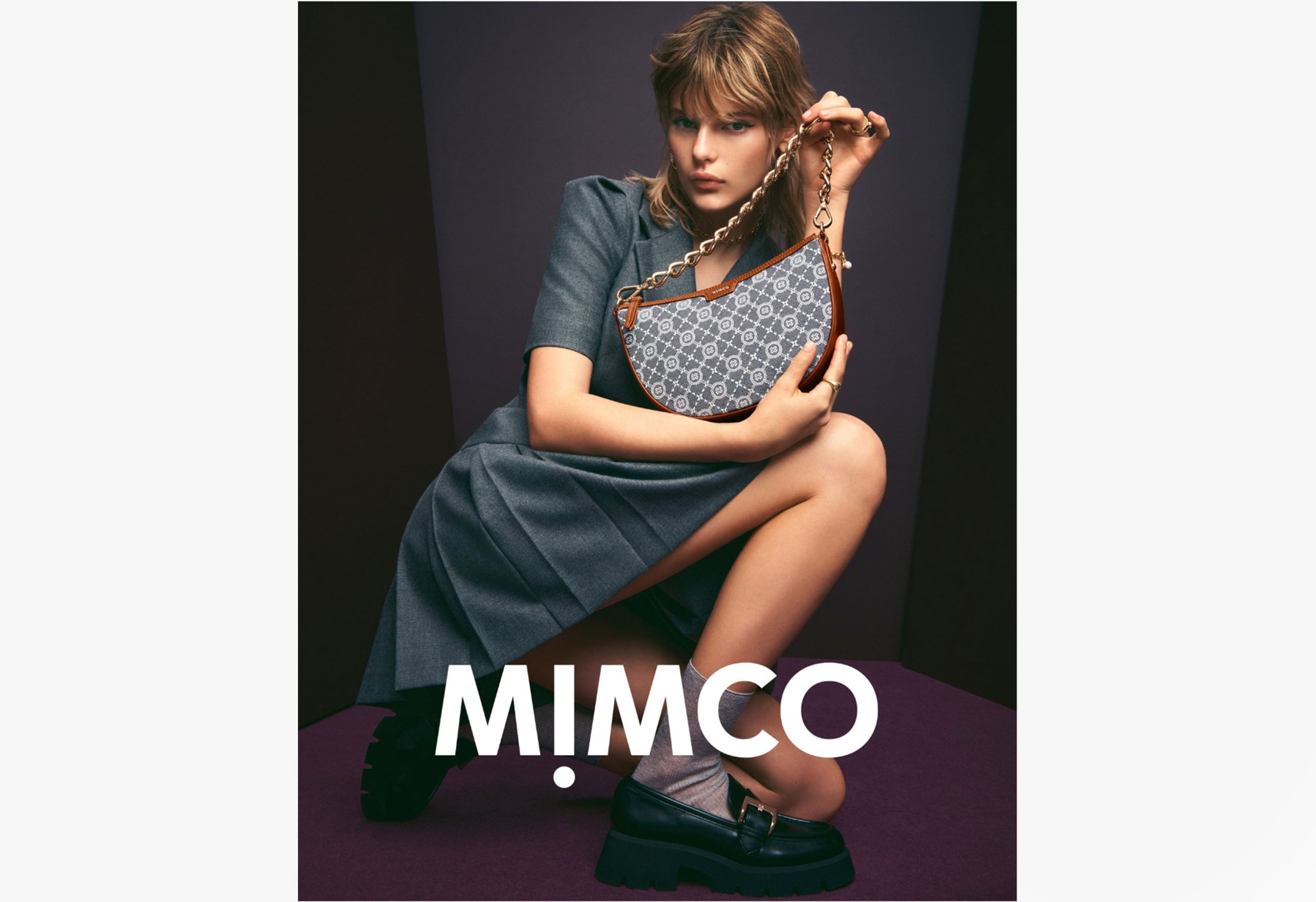 Fashion shot: woman poses with patterned Mimco handbag, wearing gray dress and platform loafers, kneeling against a dark purple backdrop.
