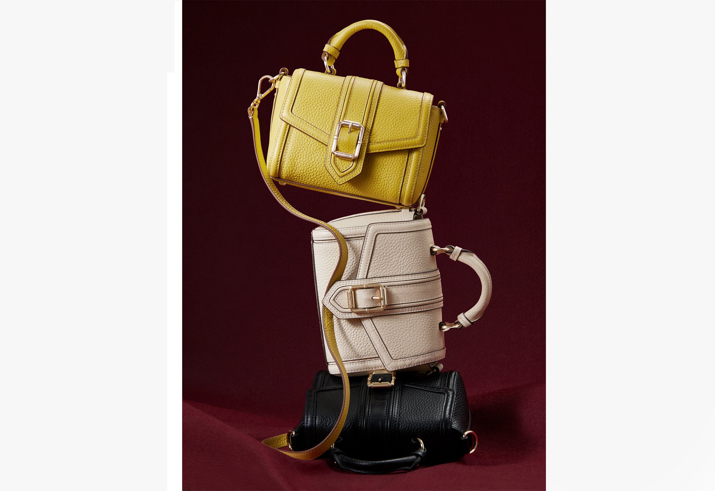Three leather handbags stacked vertically: a yellow top handle bag, a white satchel, and a black crossbody bag on burgundy fabric.