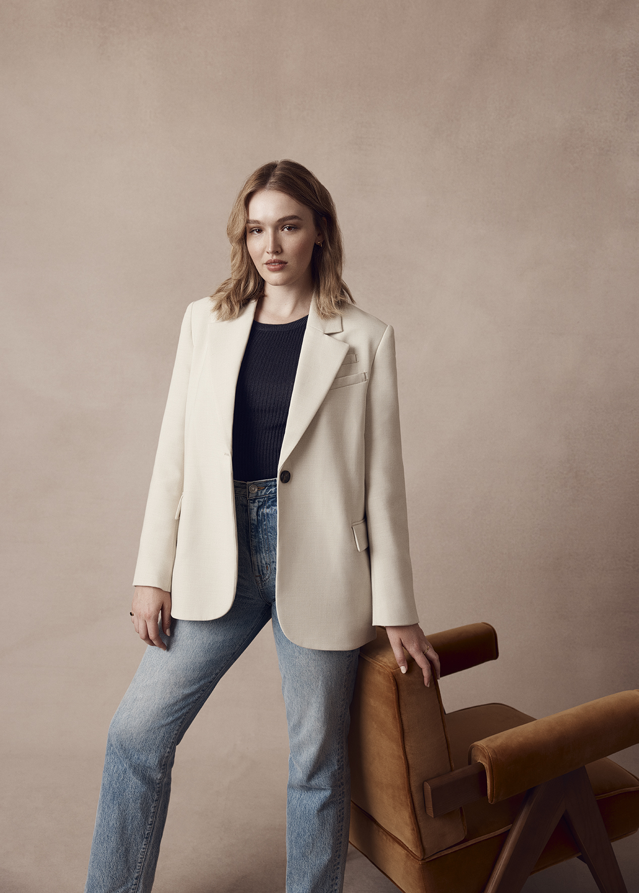 A young woman stands in a photo studio wearing a blazer and blue jeans with a modern chair visible. Neutral studio background.