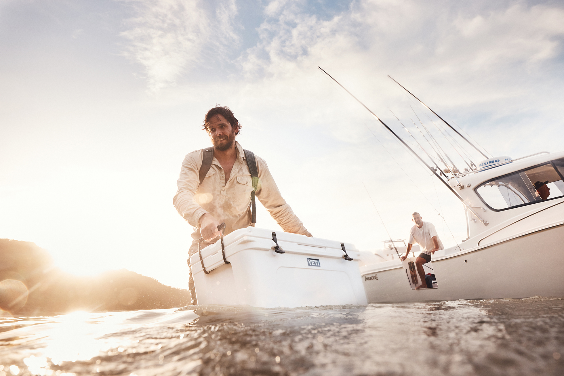 A man in water carries a white cooler toward a boat in sunlight, creating a bright, outdoor fishing or recreational scene.