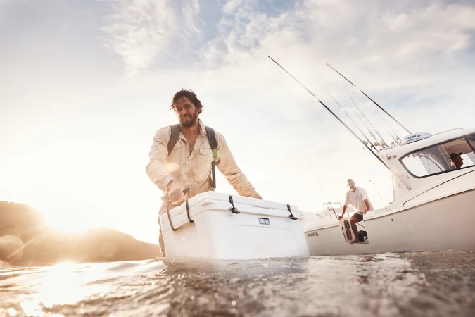 A man in water carries a white cooler toward a boat in sunlight, creating a bright, outdoor fishing or recreational scene.
