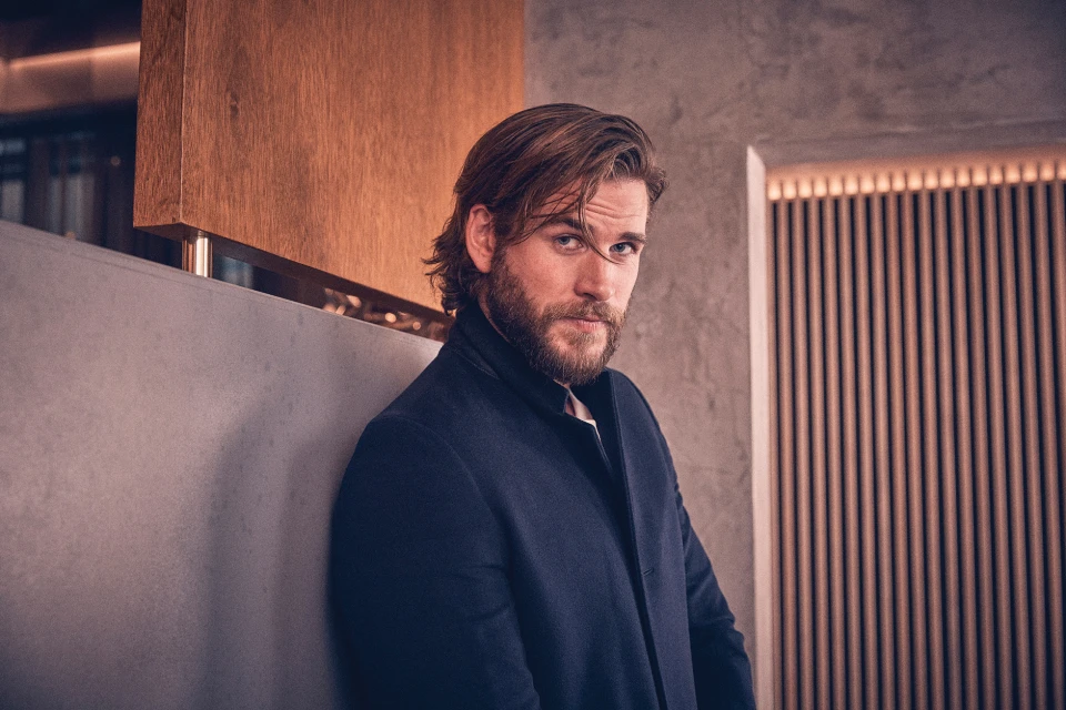 Close-up portrait of a bearded adult male with brown hair and a dark blue overcoat in front of a modern, minimalist backdrop.
