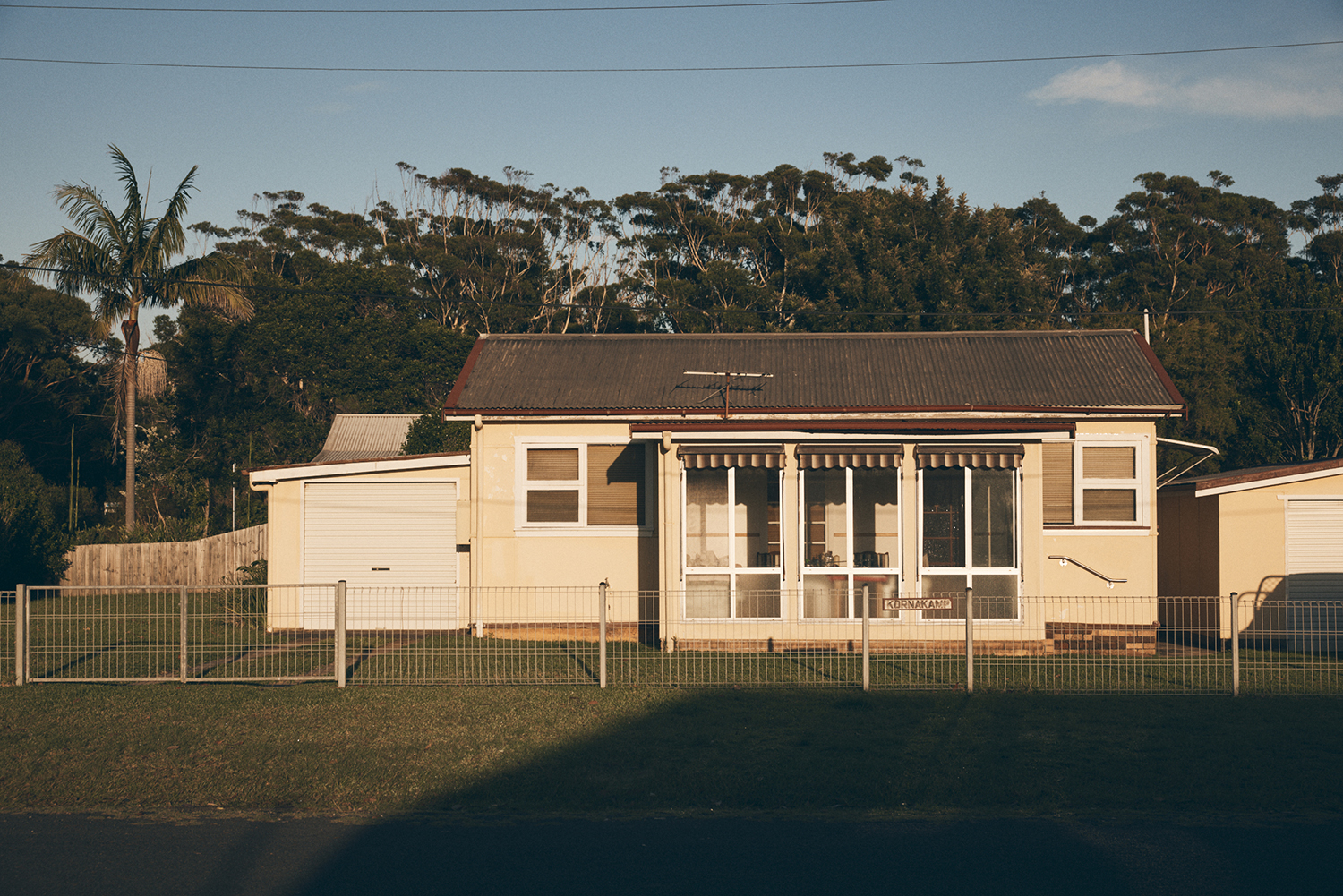 A residential building with a brown roof, white siding, and awnings over the windows. A yard and fence are visible in front of the house.