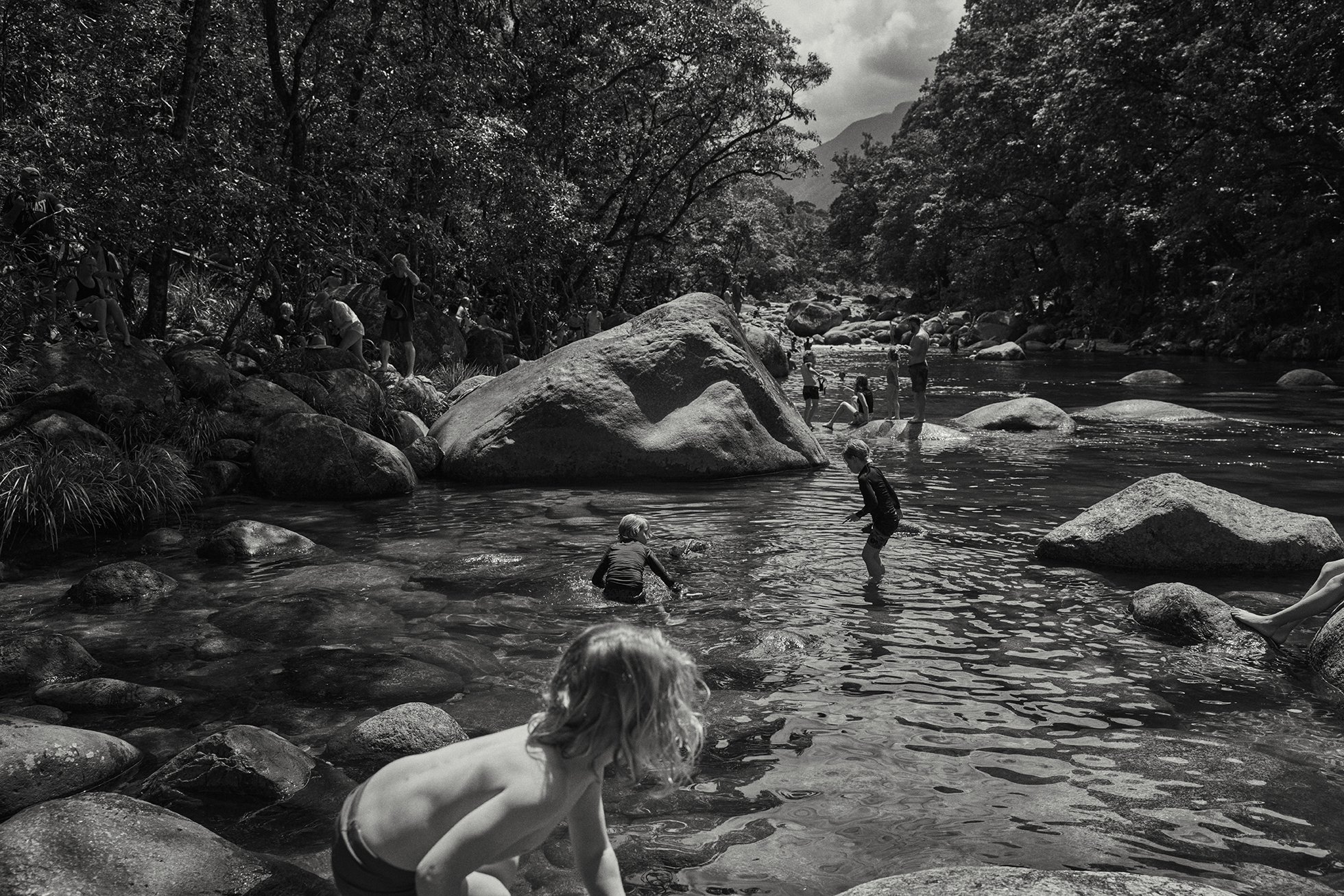 Black and white photo capturing children wading in a river surrounded by lush trees and rocks, bathed in soft, filtered sunlight.