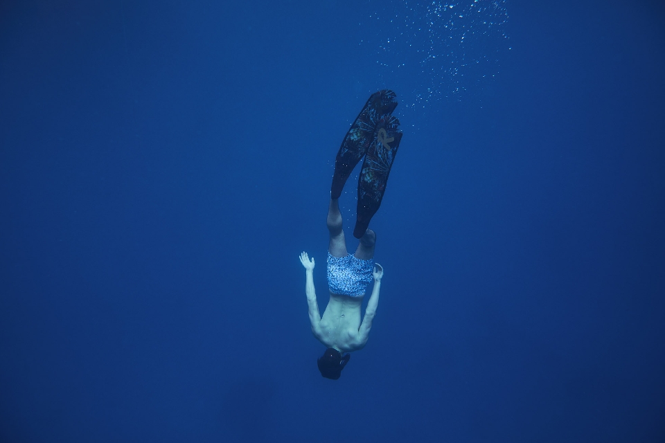 Underwater shot of a shirtless man with fins swimming down into deep blue ocean water with sunlight piercing the surface above.
