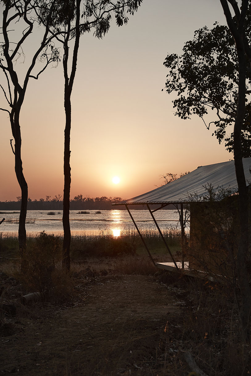 Serene sunset over a tranquil lake, framed by silhouetted trees and the corner of a simple structure, evoking peace.