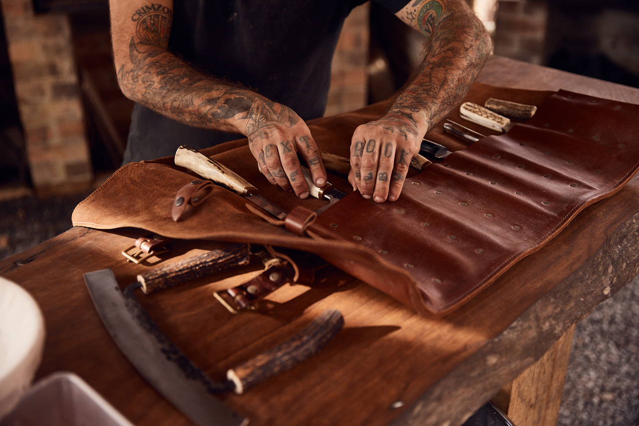 Tattooed craftsperson working with hand tools, arranged inside a leather tool roll, on a wooden workbench in dim, soft lighting.