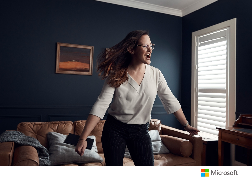 A woman with glasses, a white shirt, and dark pants dances in a room with dark blue walls, a tan leather couch, and white window blinds.