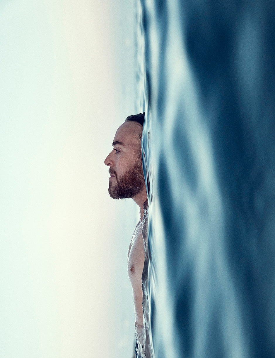 Man with a beard swimming in blue ocean water, shot in portrait orientation and side profile.