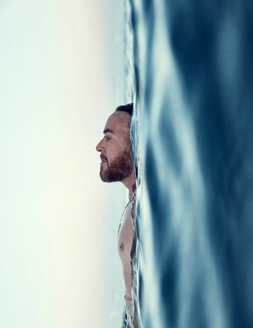Man with a beard swimming in blue ocean water, shot in portrait orientation and side profile.