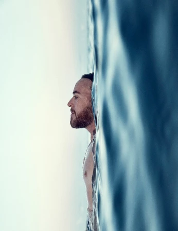 Man with a beard swimming in blue ocean water, shot in portrait orientation and side profile.