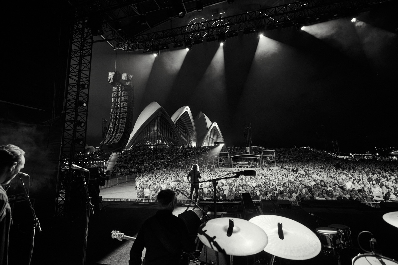 Concert view from stage shows a band performing for a large crowd with spotlights and the Sydney Opera House in the background.