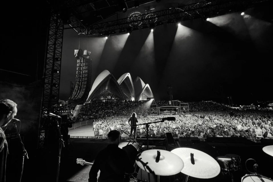 Concert view from stage shows a band performing for a large crowd with spotlights and the Sydney Opera House in the background.