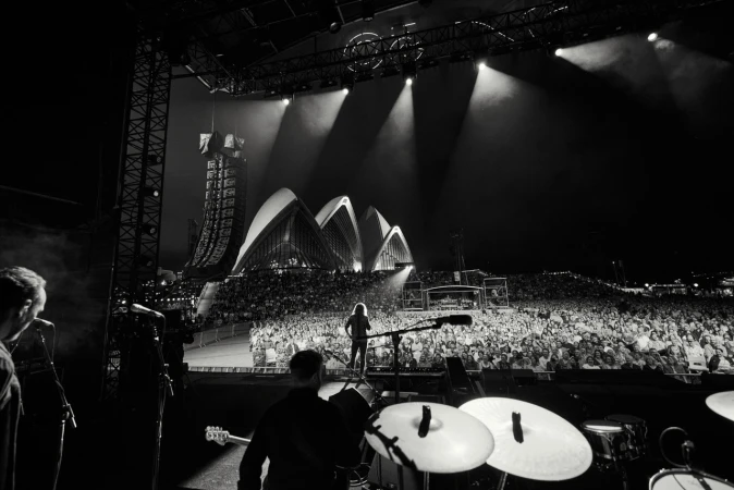 Concert view from stage shows a band performing for a large crowd with spotlights and the Sydney Opera House in the background.
