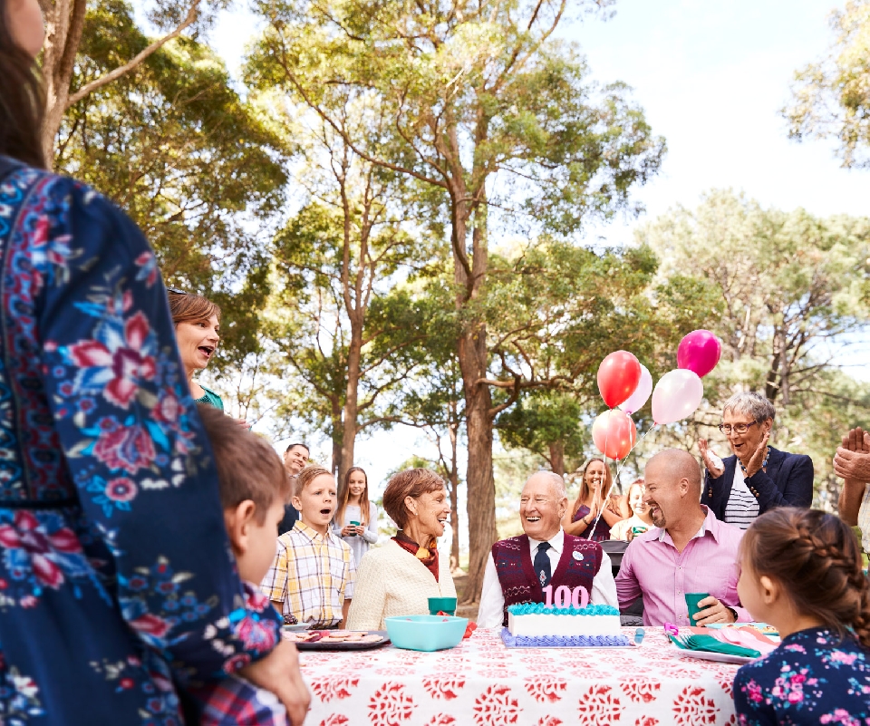 Outdoor birthday party for a centenarian with a diverse group of adults and children gathered around a table with cake and balloons.