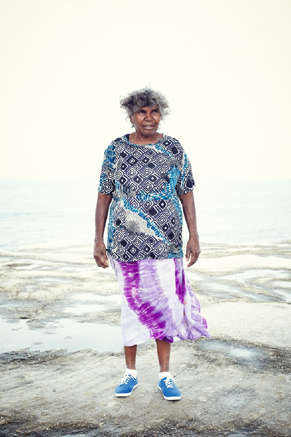 Elderly Aboriginal woman stands on a rocky outcrop near the ocean, wearing a patterned shirt, tie-dye skirt, and blue sneakers.