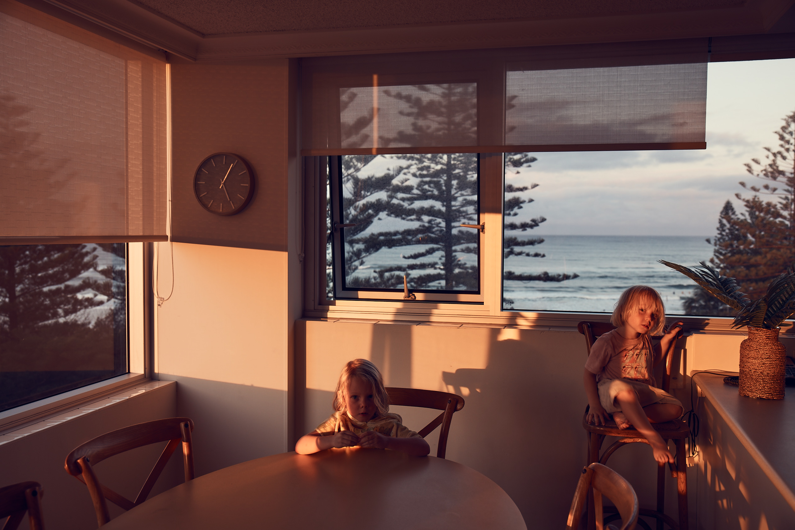 Two young children are sitting in a room with a view of the ocean through a window during sunset, casting golden light throughout the interior.