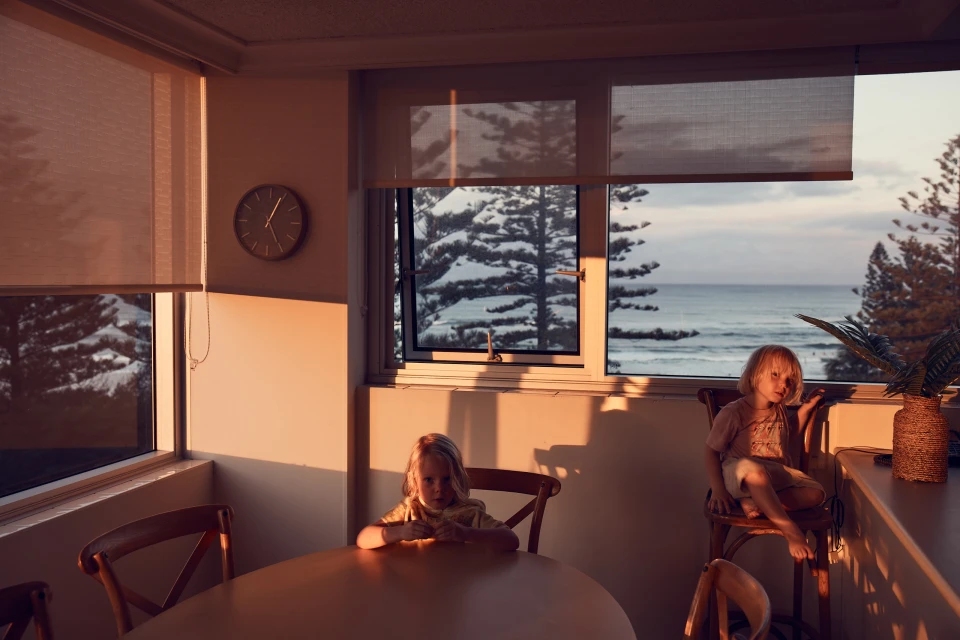 Two young children are sitting in a room with a view of the ocean through a window during sunset, casting golden light throughout the interior.
