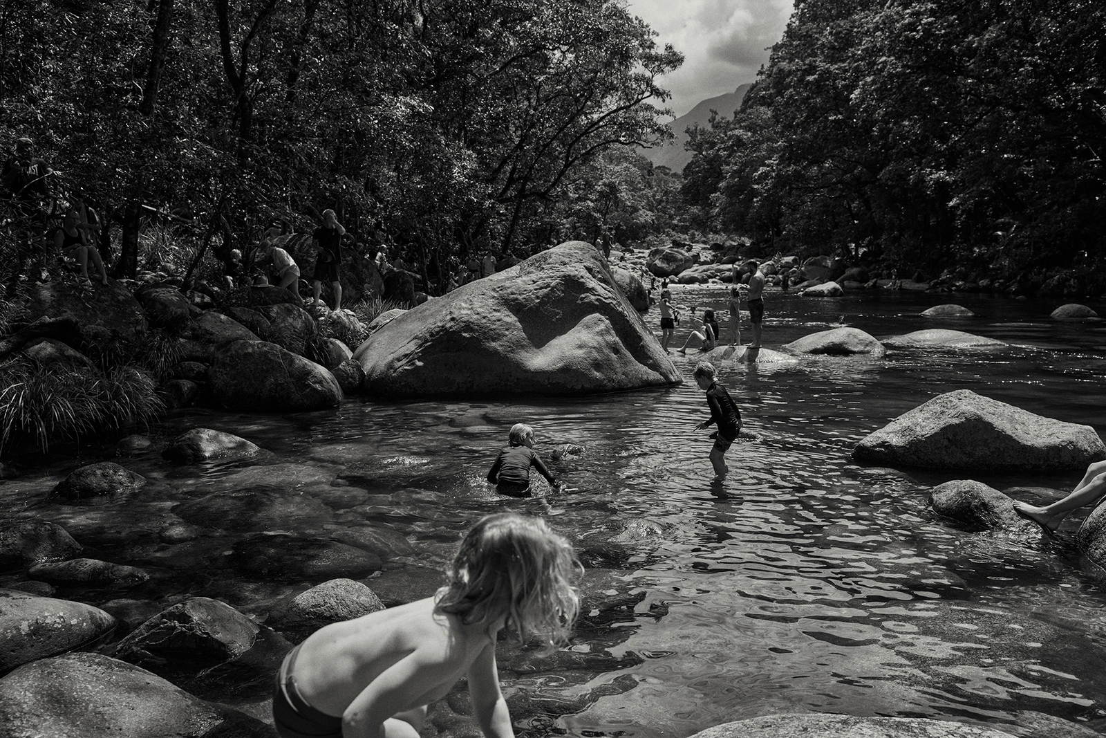 Black and white photograph by Damian Bennett of people swimming and wading in a rocky river surrounded by lush forest.