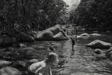 Black and white photograph by Damian Bennett of people swimming and wading in a rocky river surrounded by lush forest.