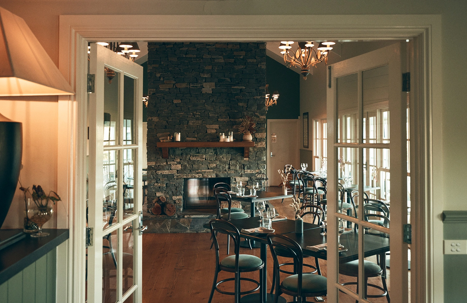 Interior view of a restaurant through open double doors, featuring a stone fireplace and wooden tables.