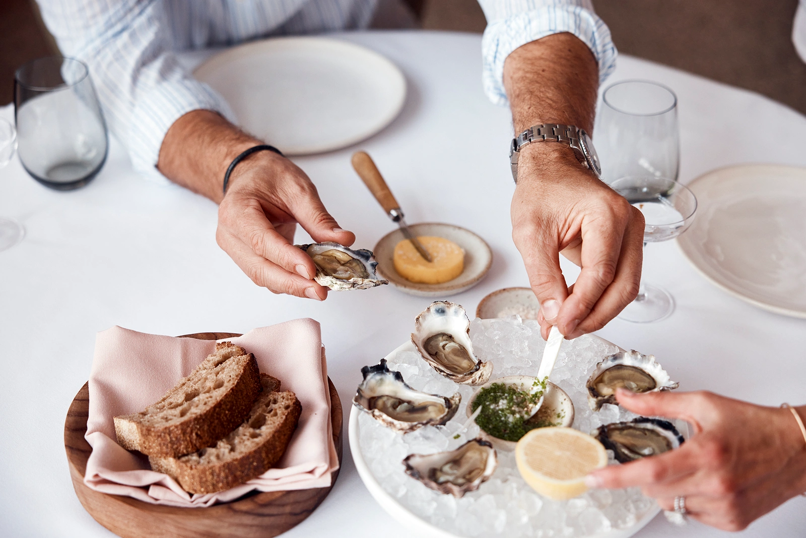 Close-up of people at a dining table holding fresh oysters on ice, bread, and condiments by Damian Bennett