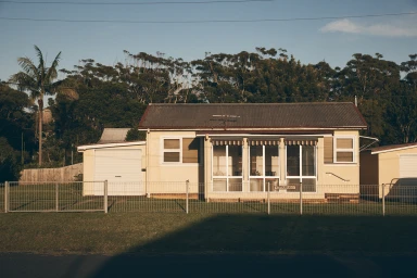 Single-story pale yellow house with dark corrugated roof behind a metal wire fence under a clear blue sky in Sydney.