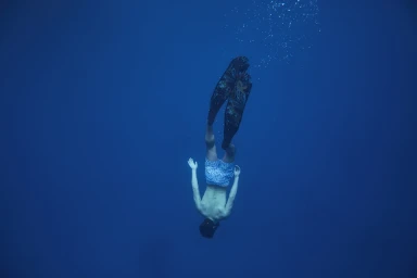A freediver wearing patterned swim shorts and long fins descending headfirst into deep blue ocean water.