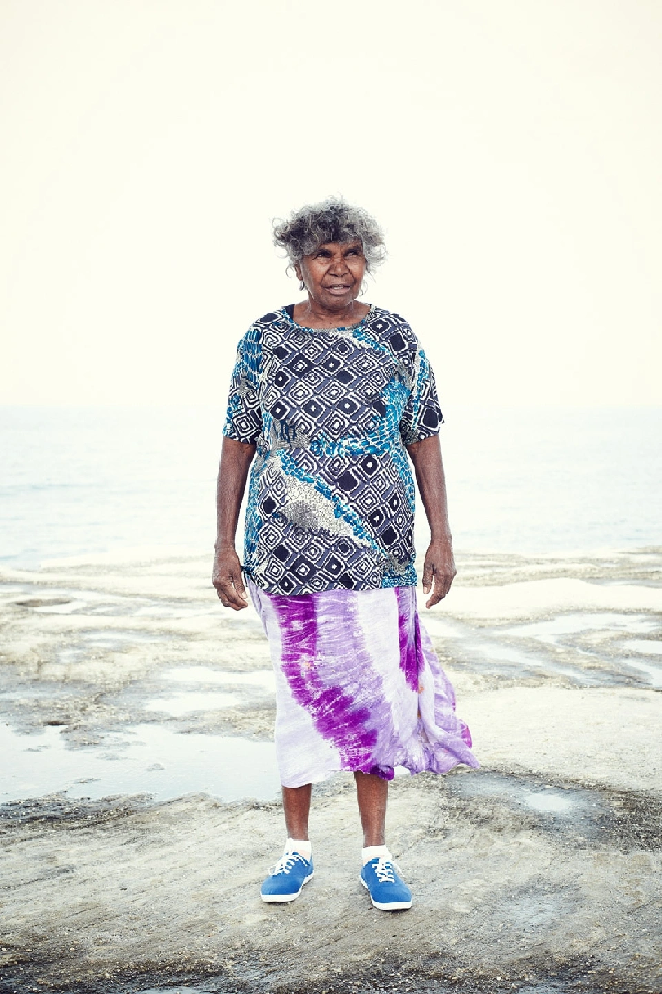 An elderly indigenous woman standing on a rocky shoreline wearing a patterned patterned top and purple skirt, photographed by Damian Bennett.