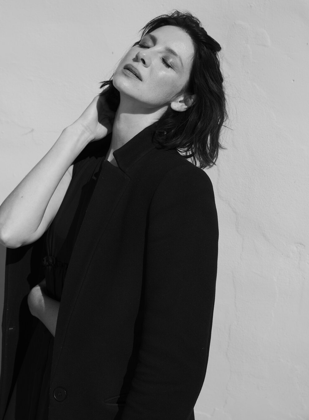 Monochrome portrait of a woman in a jacket against a textured wall, eyes closed in sunlight, with short dark hair and fair skin.
