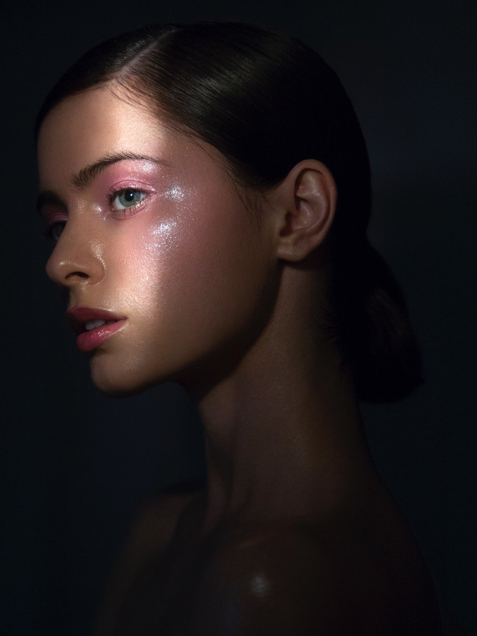 Glamour shot of a young adult woman with pink eyeshadow and glossy skin in dark studio setting with directed light.