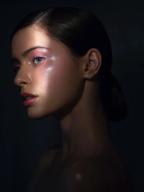 Glamour shot of a young adult woman with pink eyeshadow and glossy skin in dark studio setting with directed light.