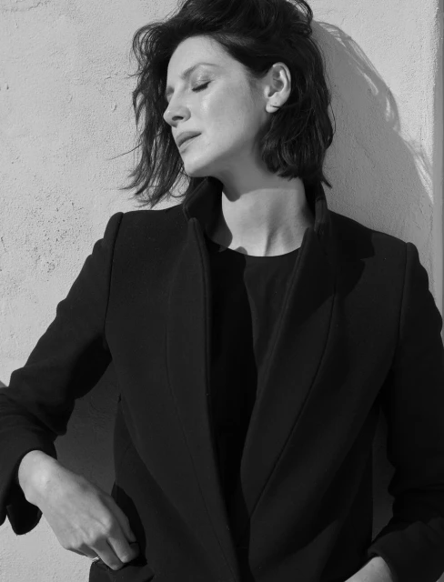 Black and white portrait of a woman with dark hair, wearing a blazer. She is in an outdoor setting against a textured wall.