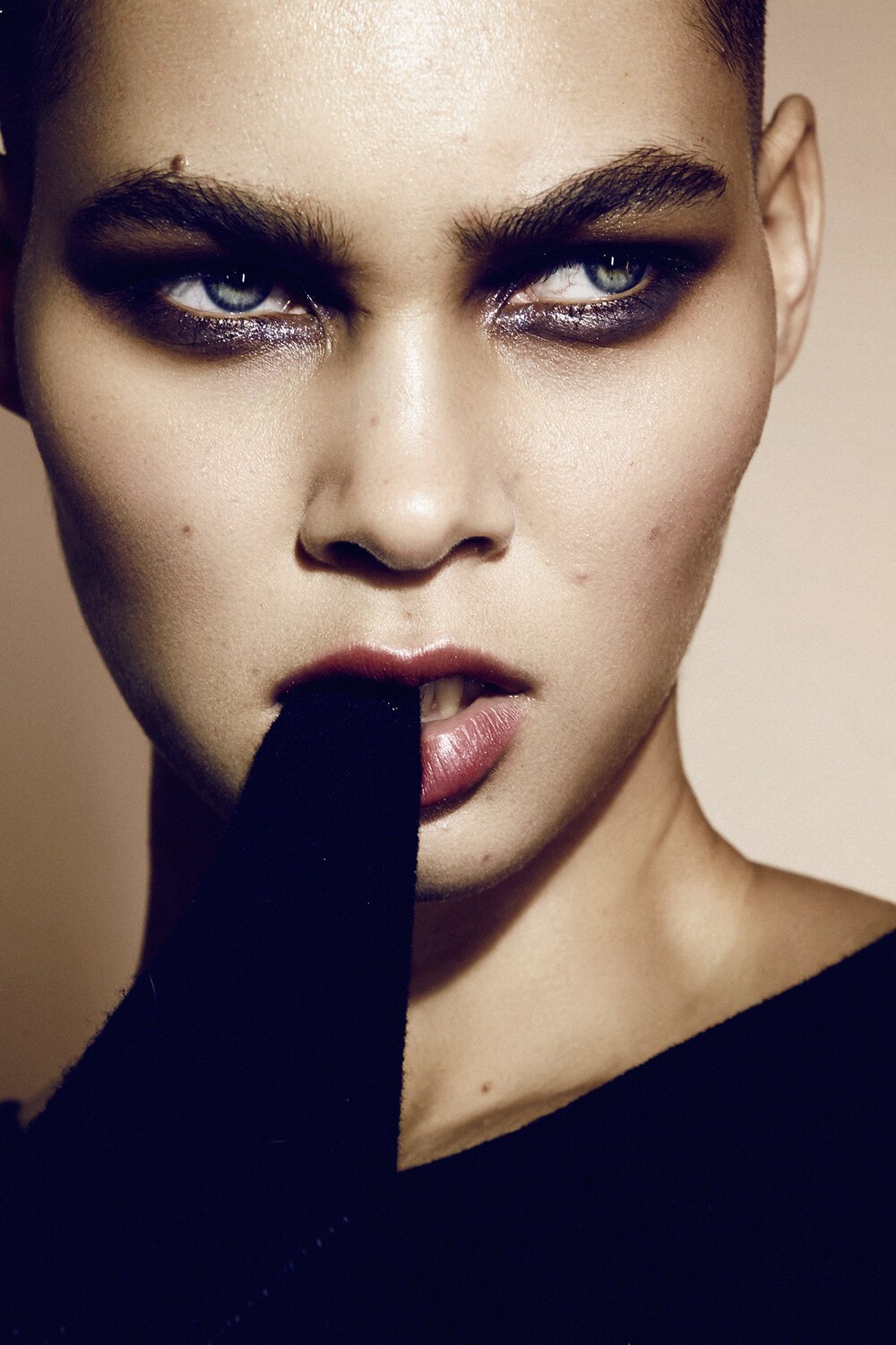 Close-up studio portrait of a woman with dramatic makeup and slicked-back short hair, wearing a black garment, against a neutral background.