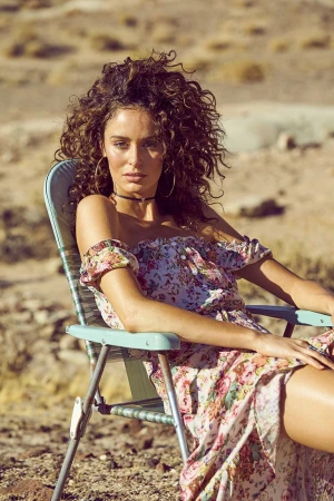 Woman with voluminous curly hair seated in a vintage folding lawn chair in an arid outdoor setting wearing a floral dress.